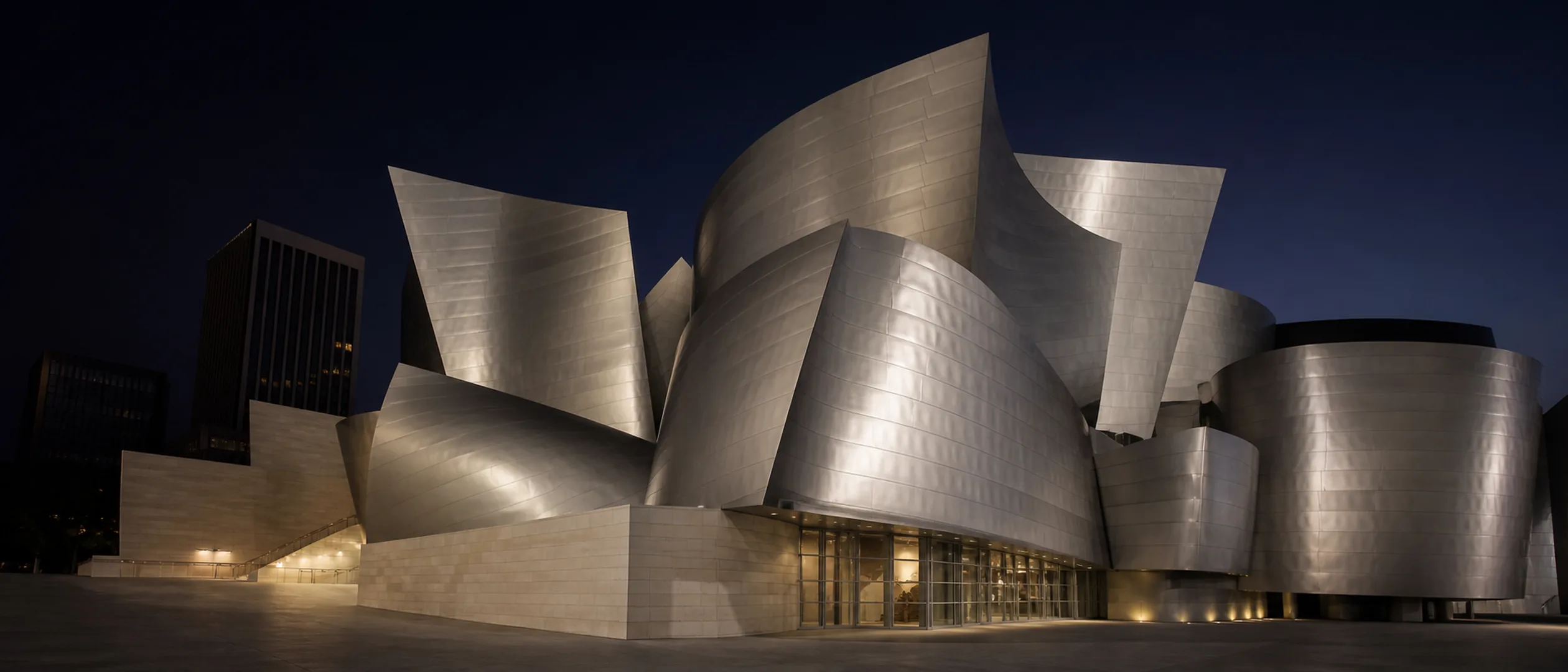 The curved stainless-steel sails of Walt Disney Concert Hall rise above a quiet plaza at blue hour.
