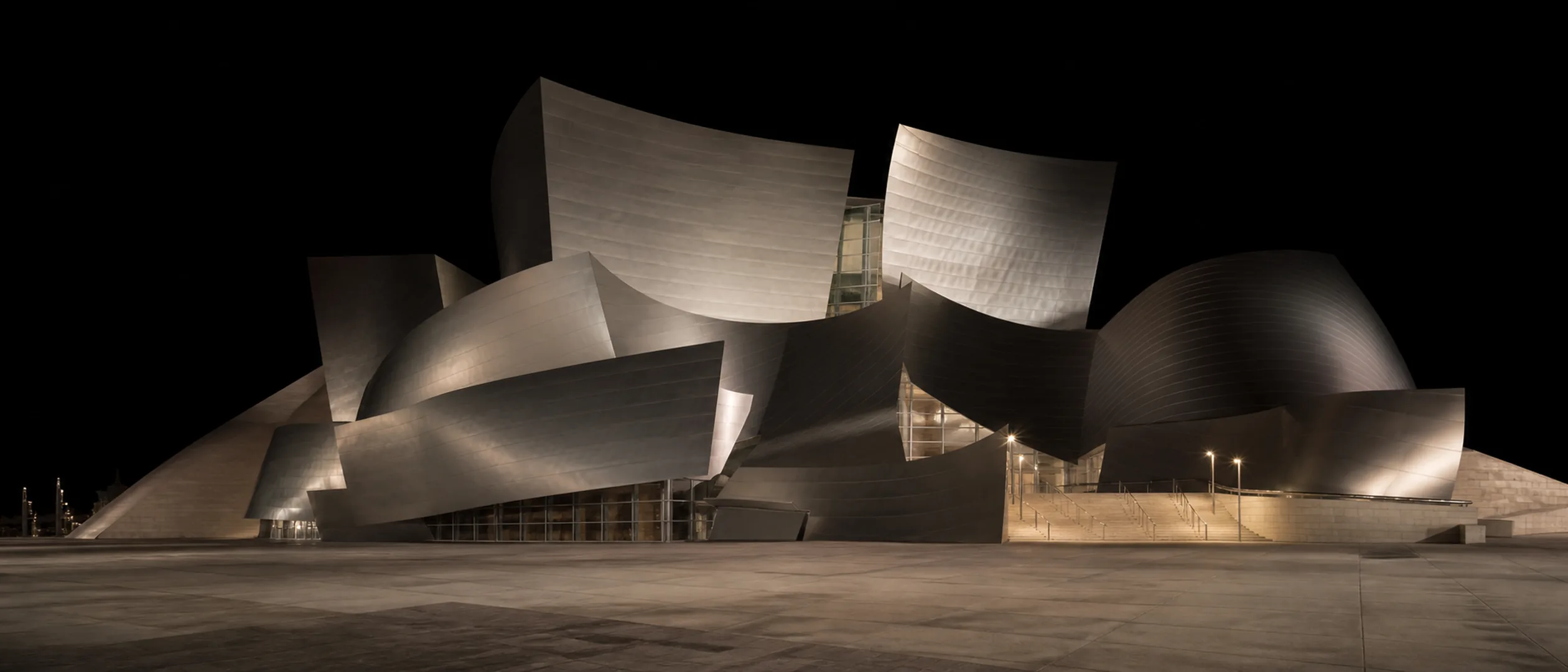 An empty stretch of Grand Avenue passes the curved stainless-steel forms of Walt Disney Concert Hall at blue hour.